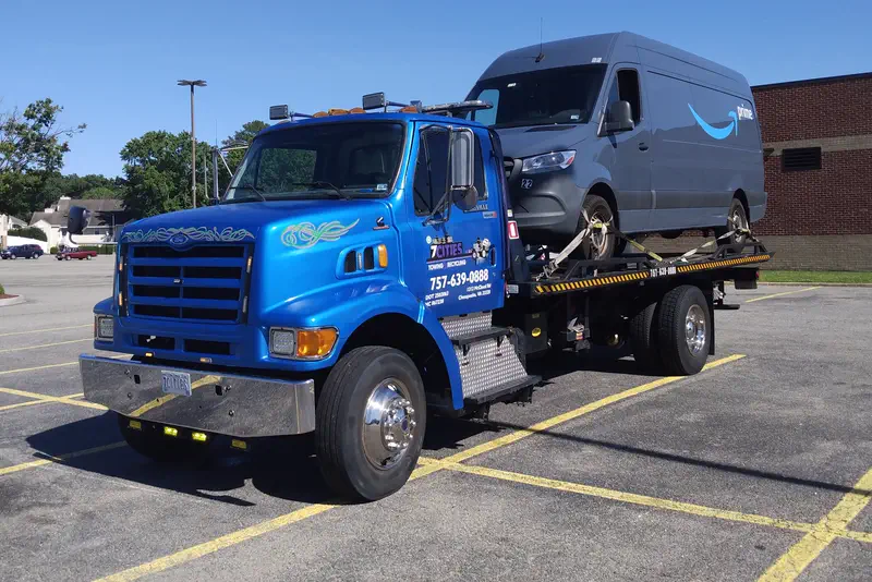 The blue 7 Cities flatbed carrying a grey delivery van across a parking lot