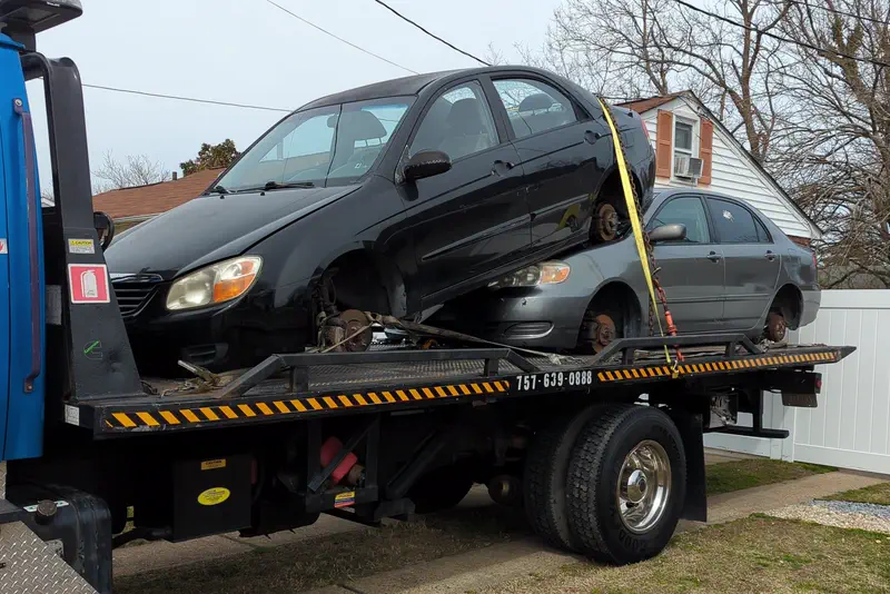 A black junk sedan with a front wheel missing, strapped down on a 7 Cities flatbed